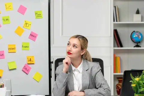 Kobieta zastanawia się nad strategią marketingową. Na tablicy karteczki z hasłami: "TEAM", "PRODUCT", "SALE", "DESIGN", "MONEY", "FINANCE", "PLAN".